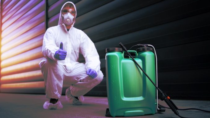 Exterminator in white protective uniform standing by reservoir with chemicals and sprayer.